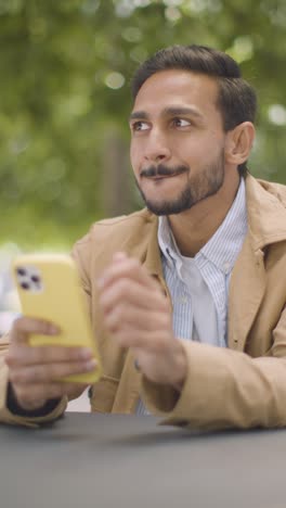 Video-Vertical-De-Un-Hombre-Musulmán-Sentado-En-Una-Mesa-Al-Aire-Libre-En-Una-Calle-De-La-Ciudad-Enviando-Mensajes-De-Texto-Hasta-La-Fecha-En-Un-Teléfono-Móvil